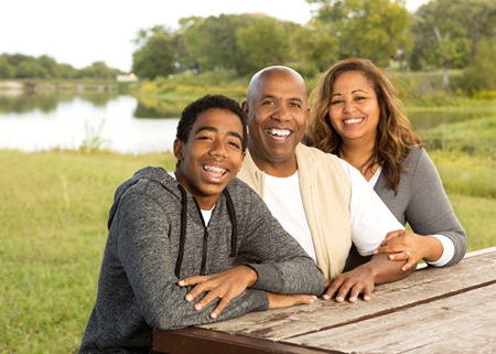 A teenage boy with braces sitting outside at a bench with his parents and smiling after seeing an orthodontist in Denison