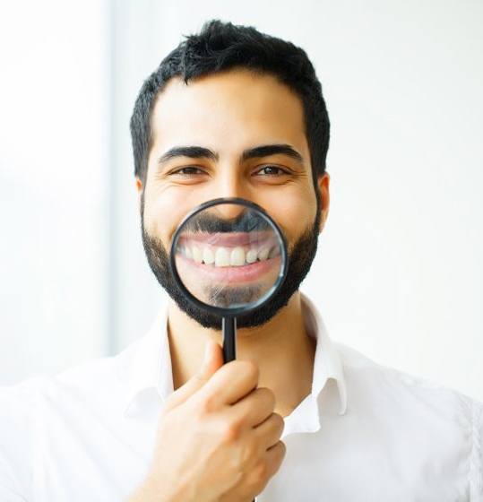 man in white shirt holding magnifying glass to his smile after porcelain veneers